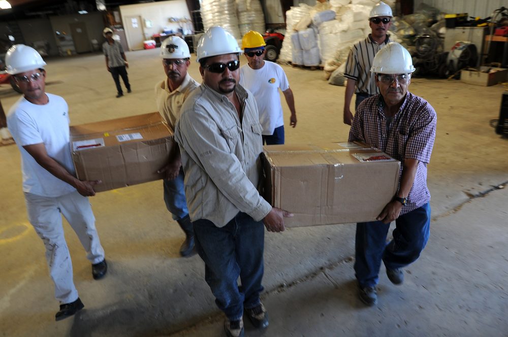A crew of environmental services contractors in hard hats and work clothes carries cardboard boxes of cleanup supplies through a staging warehouse stacked with sorbent material in the background.