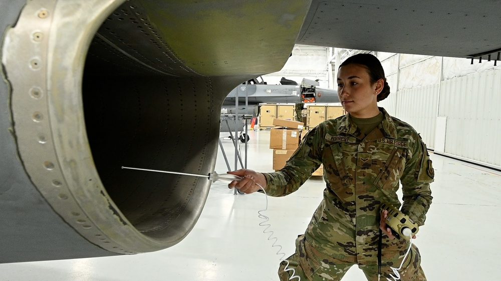 An Airman uses a 4-gas meter with a sampling probe to measure the atmosphere inside an aircraft engine intake. She holds the meter in one hand and the probe in the other, the probe extending into the confined space before any worker enters.