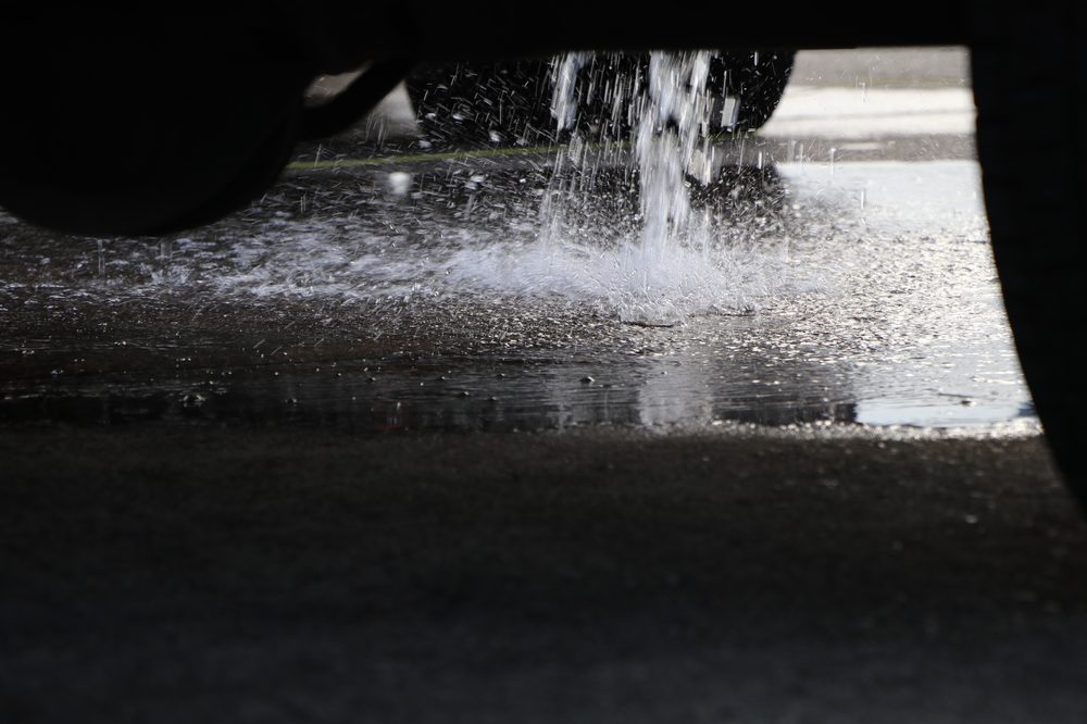 Liquid splashes from underneath a vehicle onto pavement, spreading across the ground in a small puddle. The surface is wet with the discharge.