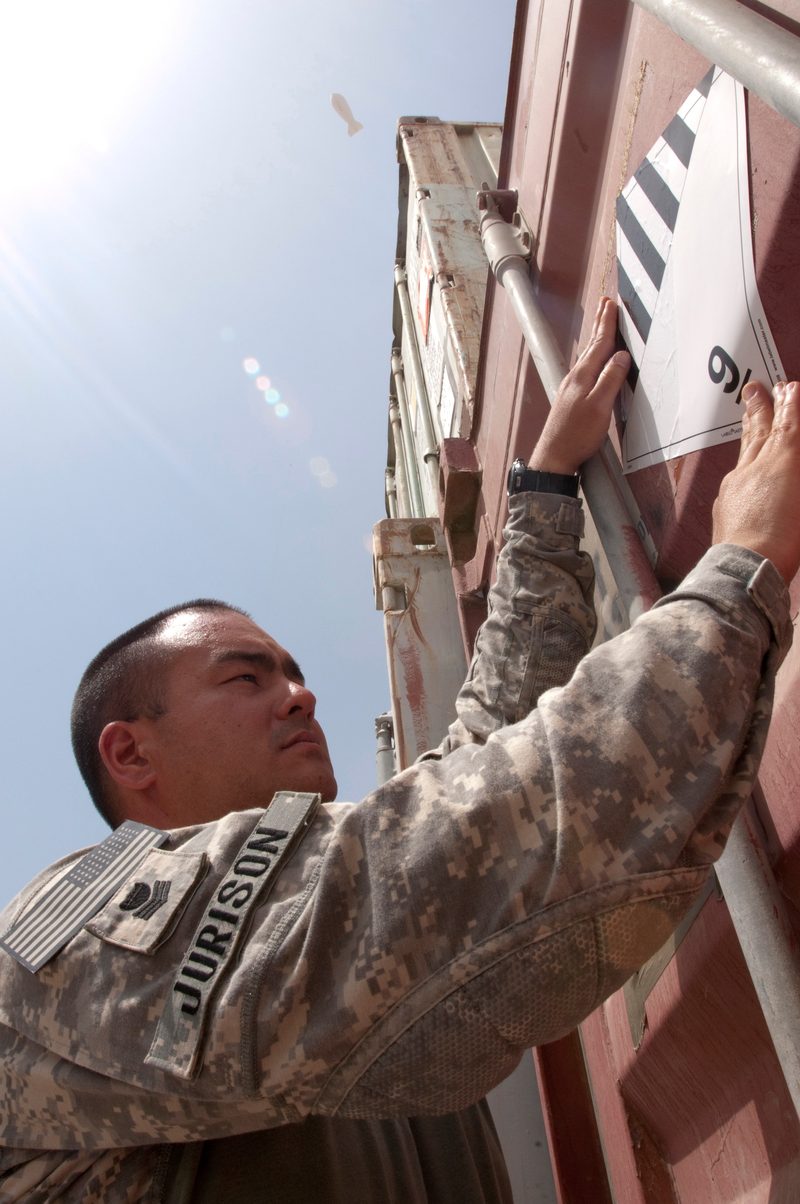A military servicemember in uniform applies a hazmat placard to the side of a shipping container, with the placard's diagonal stripe pattern visible. The number 9 indicates Class 9, Miscellaneous Hazardous Materials.