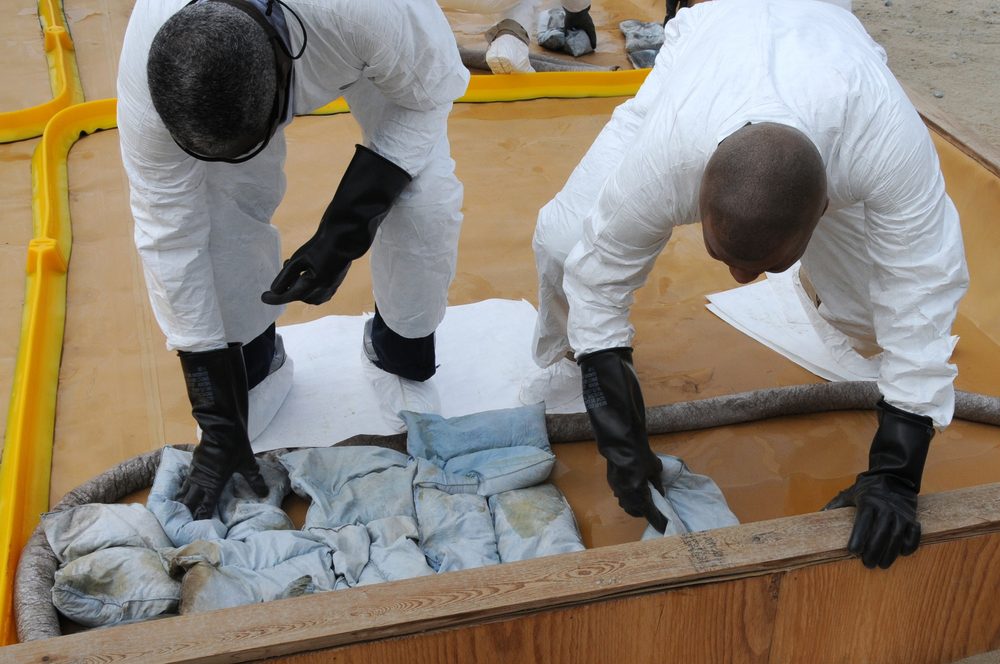 Two responders in white Tyvek suits and black neoprene gloves and boots use absorbent socks to contain a contained area and absorbent pillows to soak up the contaminated liquid inside. The absorbent pads are saturated with the spilled material.