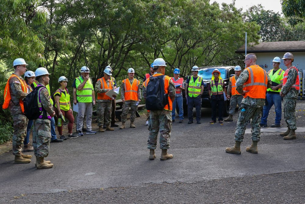 A group of uniformed military personnel and civilian inspectors in hard hats and high-visibility vests stands in a semicircle outside a regulated facility, holding clipboards and reviewing notes during a pre-walk briefing.