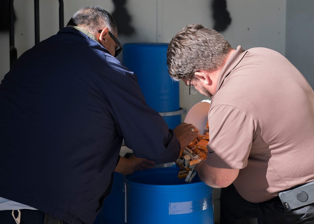 Two civilian environmental program technicians lean over a 55-gallon blue plastic drum, loading used alkaline batteries with their terminals taped for safe transport.