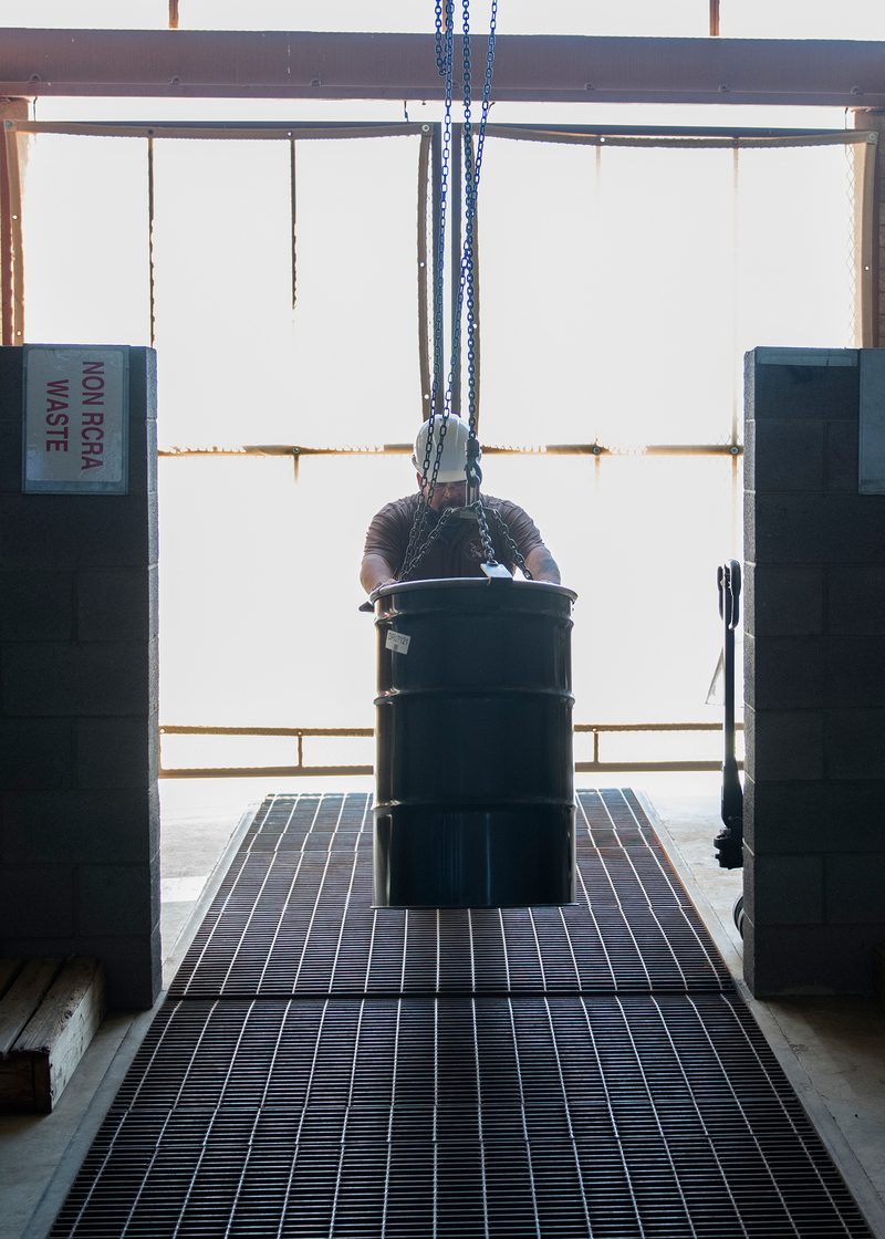 A worker in a hard hat moves a 55-gallon steel drum onto grated secondary containment flooring. A 'NON RCRA WASTE' sign is visible on the wall, indicating the area is segregated by waste classification.