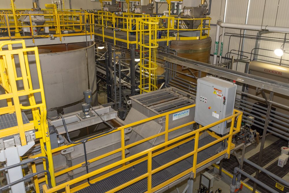 Interior of an industrial wastewater treatment plant showing clarifier tanks, treatment vessels, yellow safety railings around process equipment, and an electrical control panel.