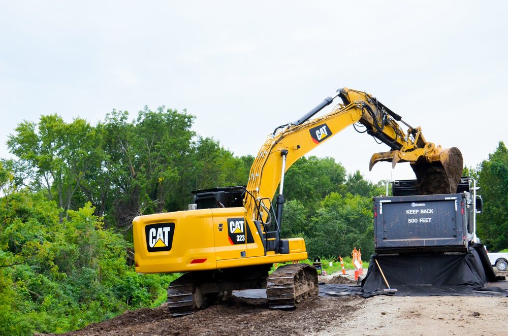 A tracked excavator loads contaminated soil from an excavation into a covered dump truck. The truck has 'KEEP BACK 500 FEET' marked on the back, and black plastic sheeting is laid out on the ground around the loading area.
