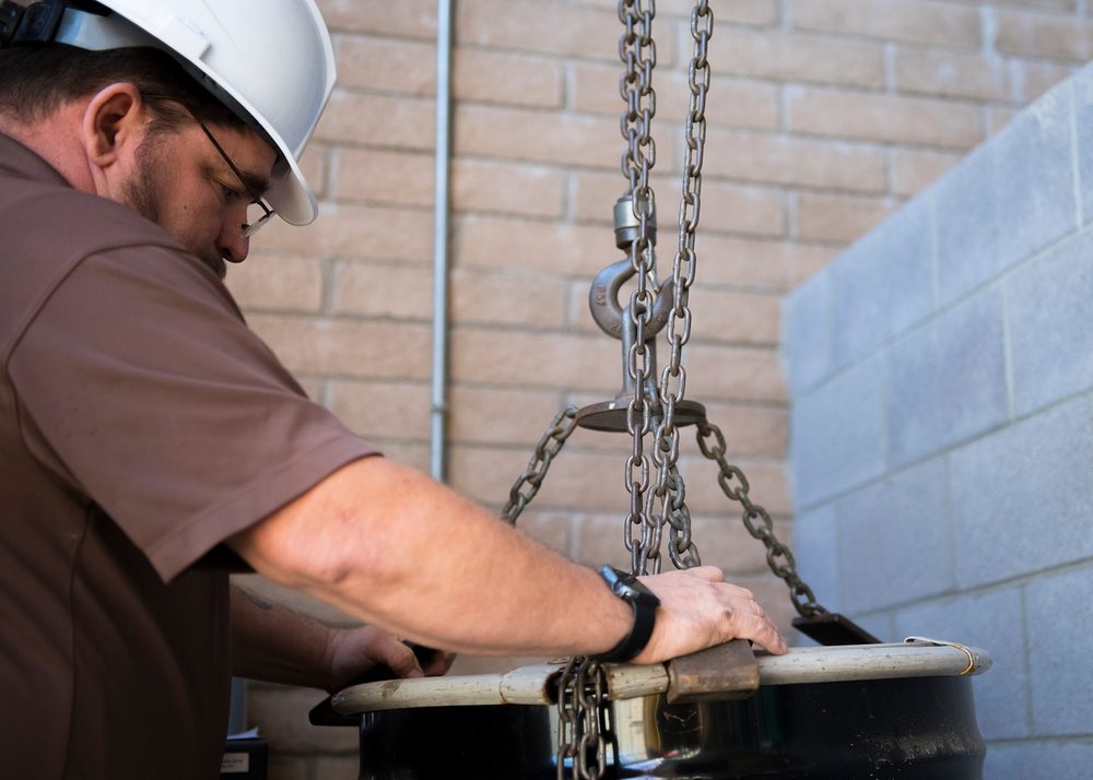 An environmental program technician in a hard hat leans over a hazardous material drum, working with a chain hoist rigged through the drum's handles for lifting.