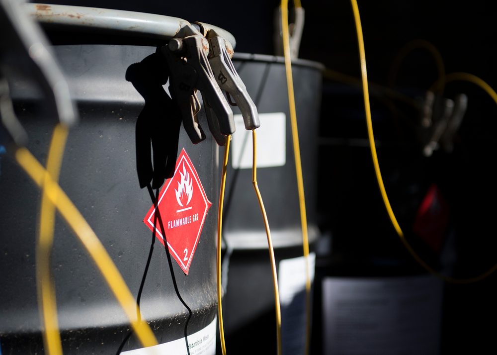 Close-up of a steel drum in a hazardous material holding facility, marked with a red Class 2 Flammable Gas placard and connected to grounding clamps and bonding wires.