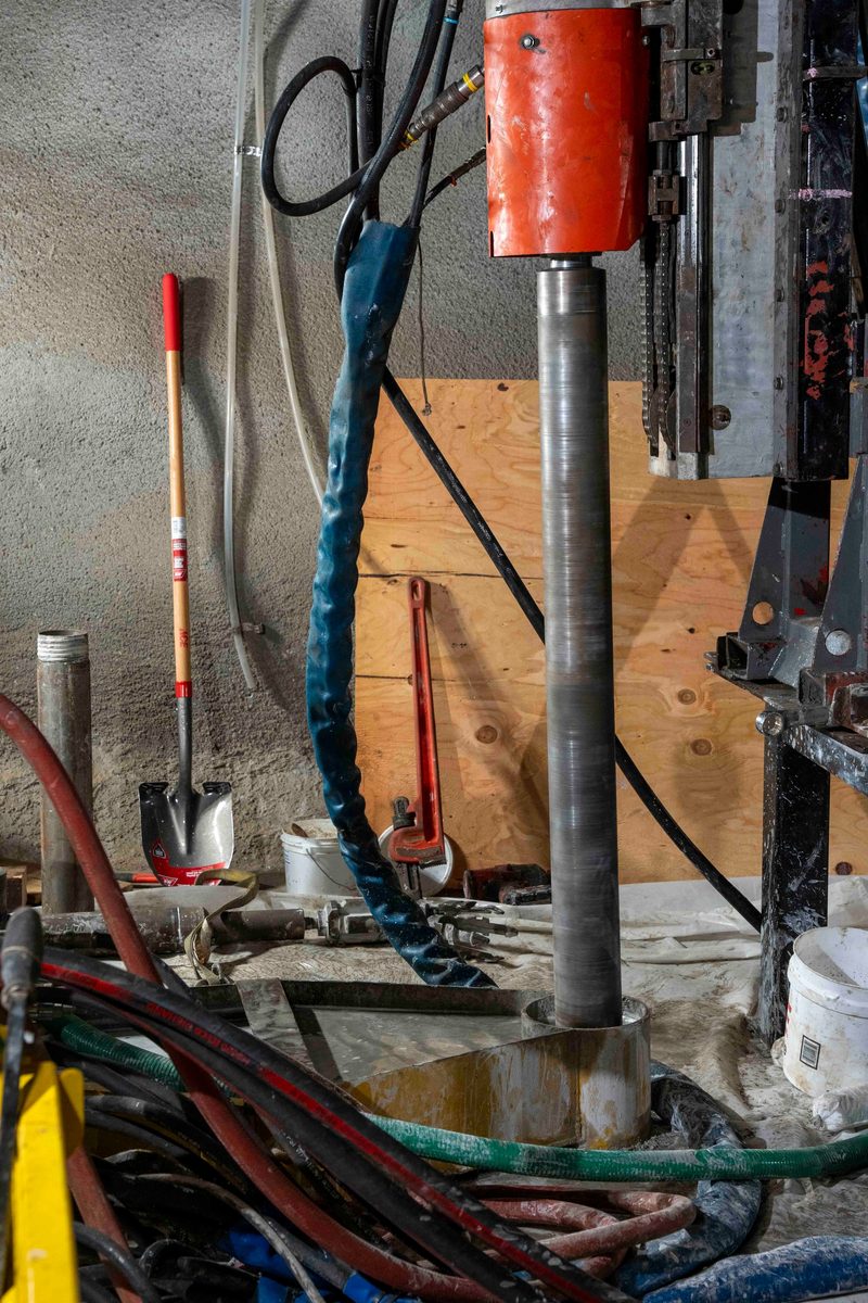 A soil coring drill rig operates inside a workspace with hoses, drilling fluid lines, a shovel, and a pipe wrench staged nearby.