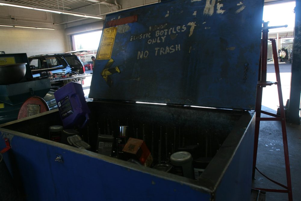 Open metal disposal bin in a vehicle maintenance shop with a hand-painted sign reading PLASTIC BOTTLES ONLY, NO TRASH on the lid, containing used oil bottles. A 55-gallon drum is visible to the left.