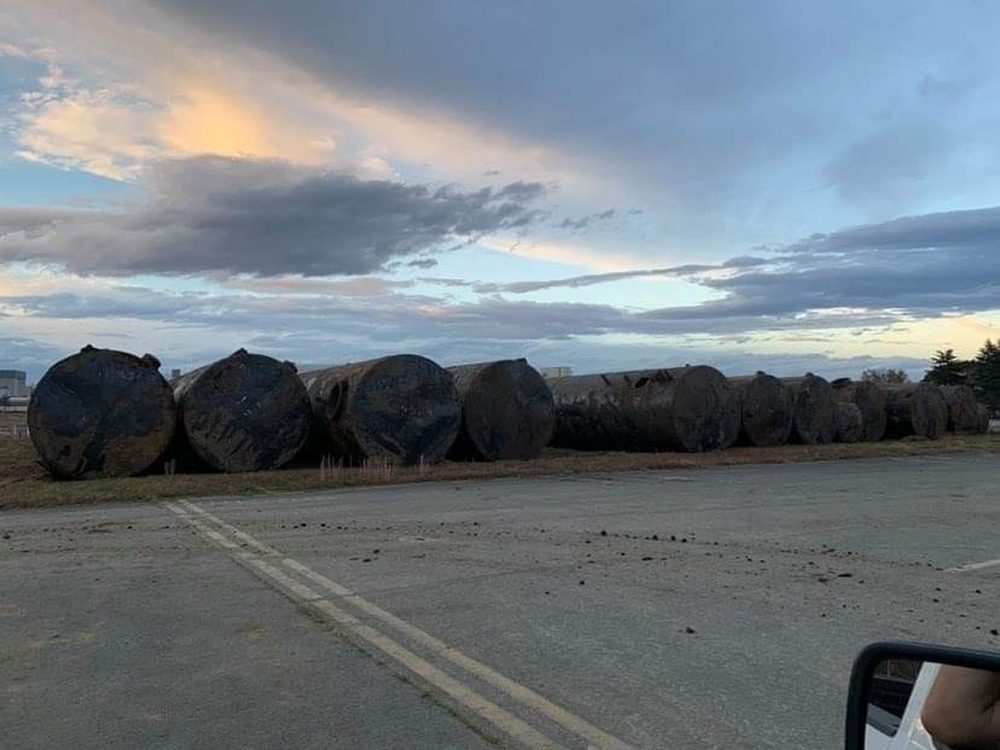 Sixteen large cylindrical underground fuel tanks lined up after excavation, showing weathering and rust on their exteriors. Each tank is roughly 12 feet in diameter.