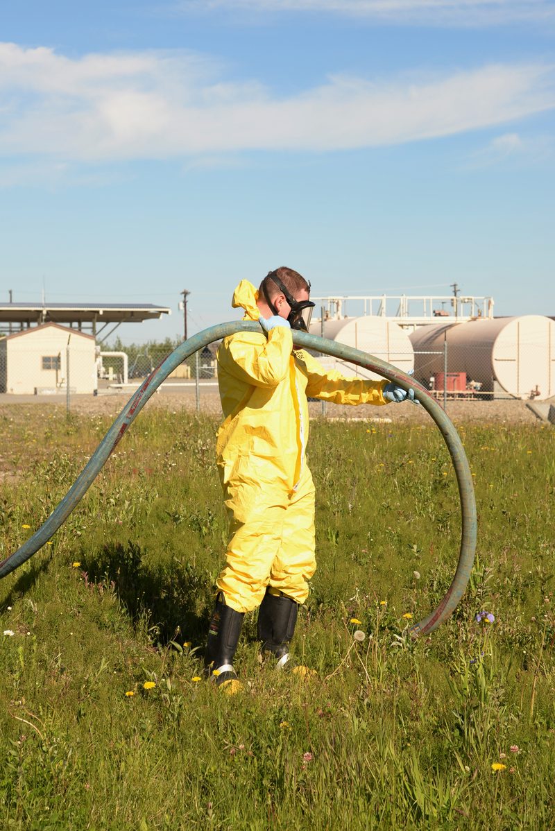 A worker in full yellow Tyvek chemical suit, respirator, and gloves carries a large vacuum hose across a field during a fuel spill response exercise. Bulk fuel storage tanks are visible in the background.