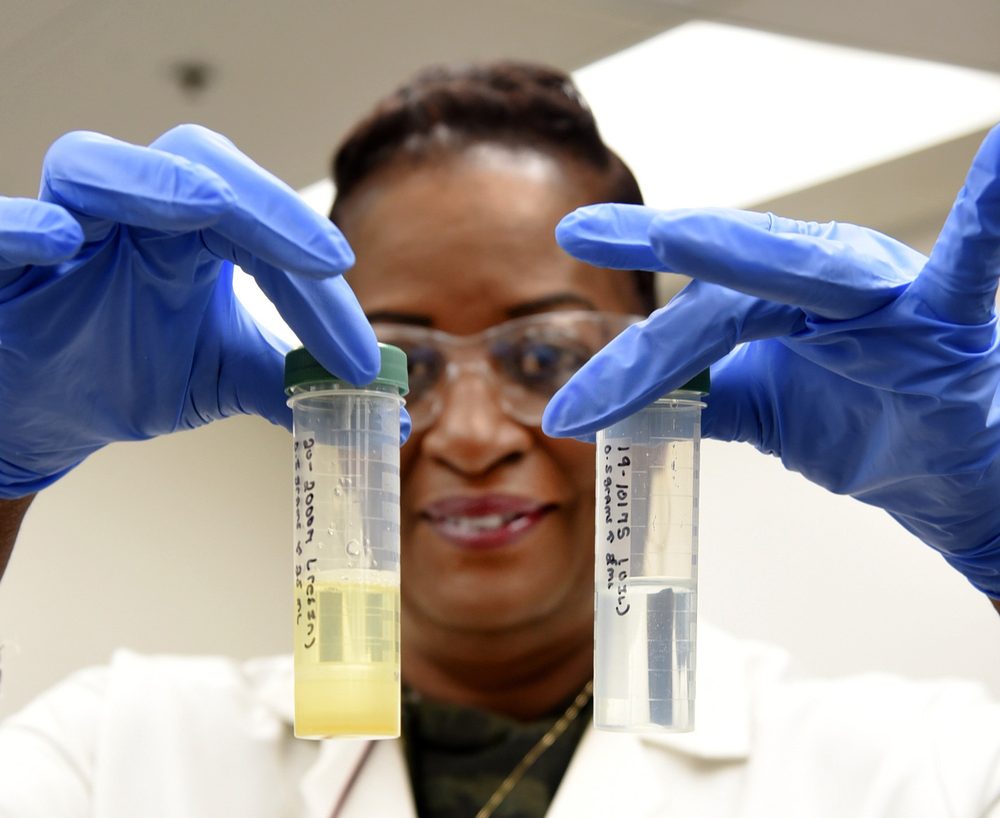 An environmental lab technician in nitrile gloves, lab coat, and safety glasses holds two labeled sample vials up to the light. Each vial bears a hand-written sample ID and date in marker.