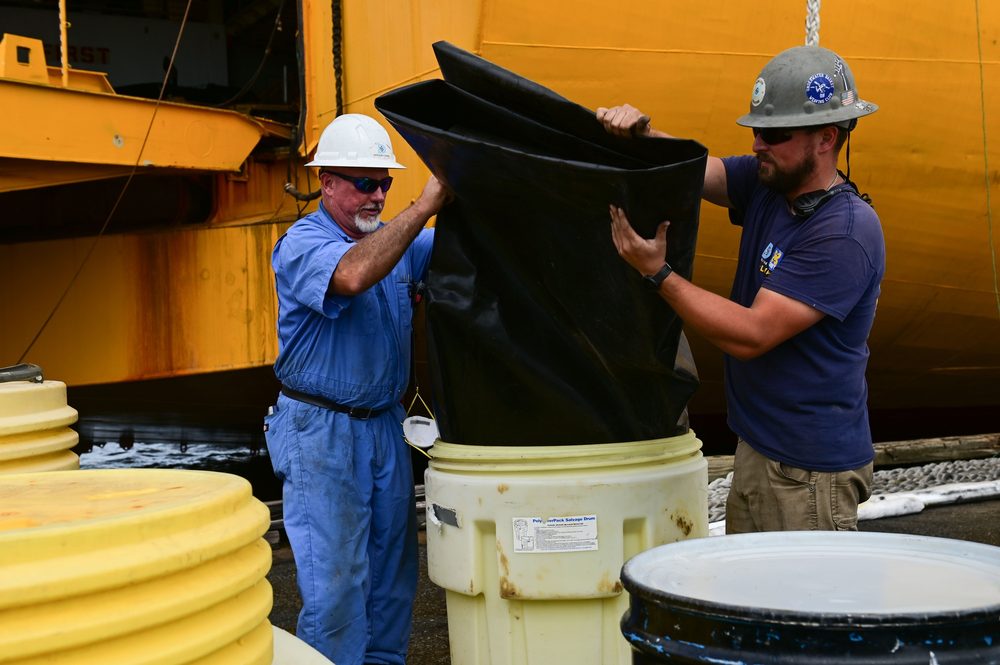 Two responders stage a portable spill containment berm at a port facility, with multiple yellow polyethylene overpack drums positioned for waste collection.