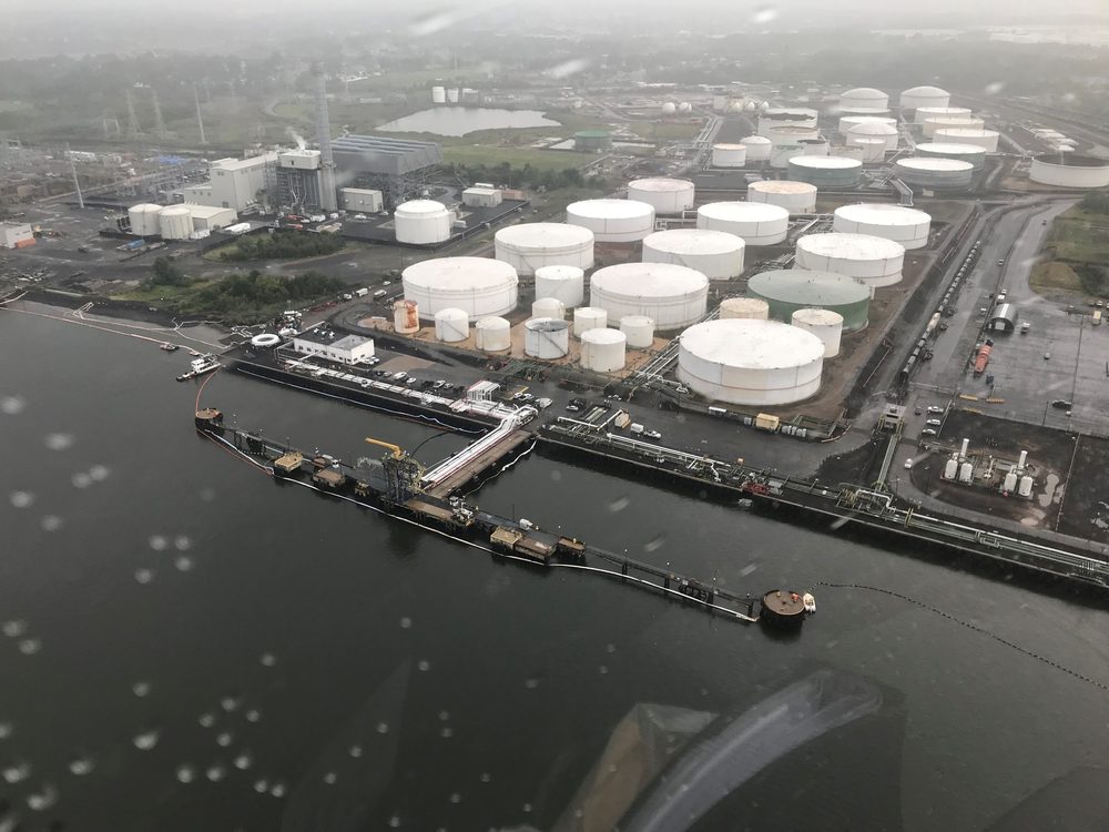 Aerial view of a large petroleum bulk storage terminal showing approximately thirty white aboveground storage tanks inside dike compounds, with a dock and tanker barge along an adjacent waterway.