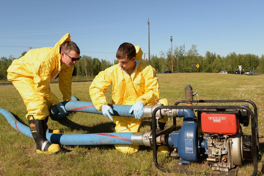 Two responders in yellow Tyvek chemical suits connect blue hoses to a diesel-powered vacuum pump in a grass field.