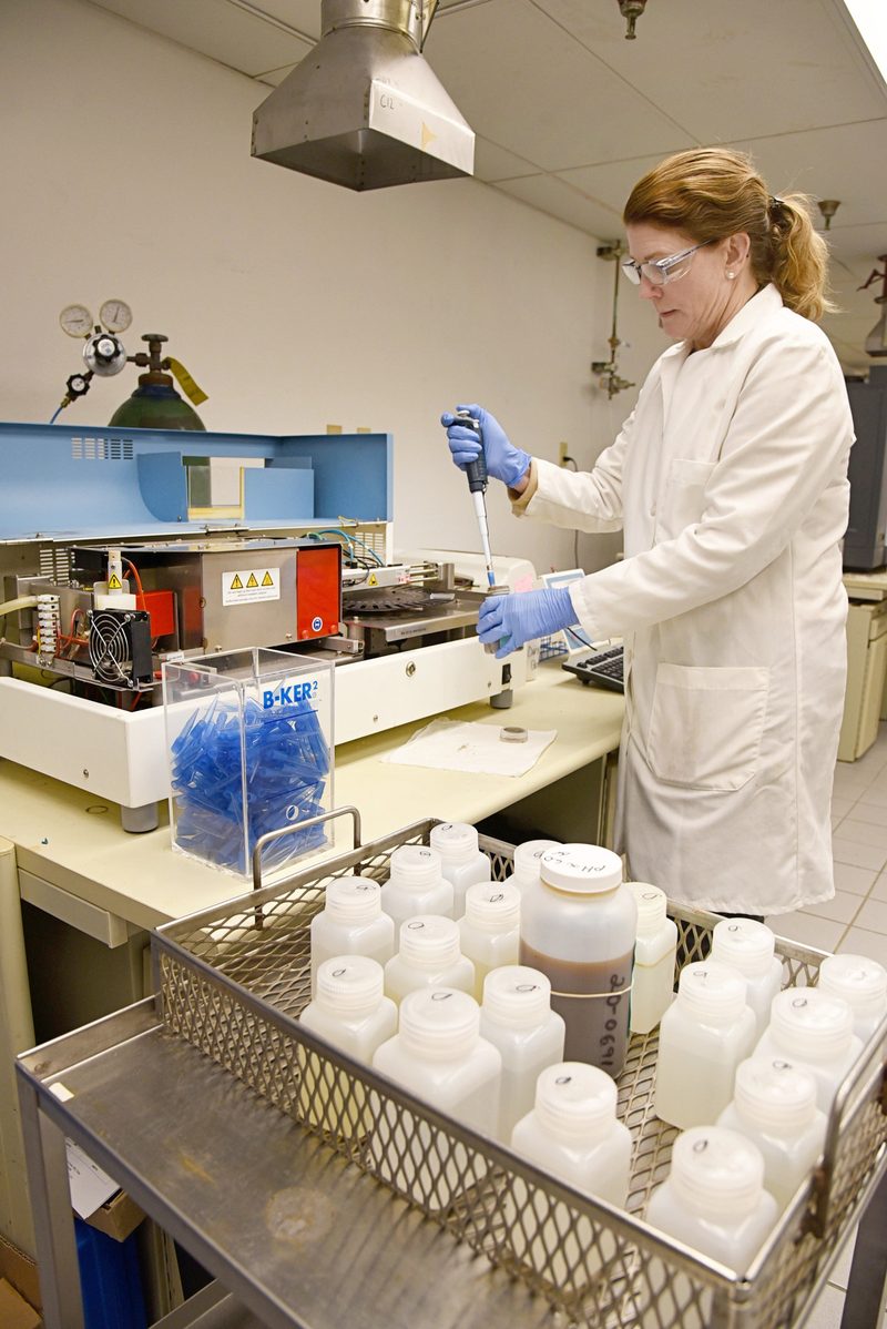 Environmental lab chemist in white coat and nitrile gloves uses a pipette to transfer a sample at a benchtop instrument, with a stainless steel basket of HDPE sample bottles in the foreground.